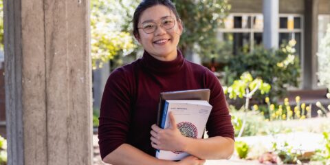 Curtin student holding study books to chest, staring directly at camera smiling, outside on Perth campus.
