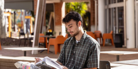 Curtin student studies at cafe tabel with book in hand on Perth campus.