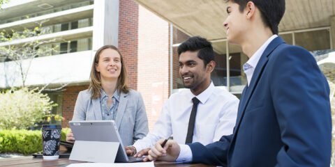 Three students sit around in business clothes looking at an iPad.