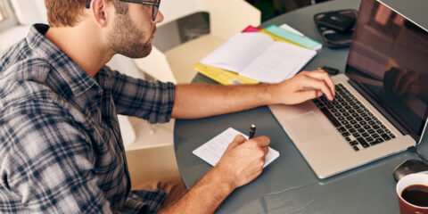 A student uses their left hand to browse their laptop and right hand to write down on a piece of paper.