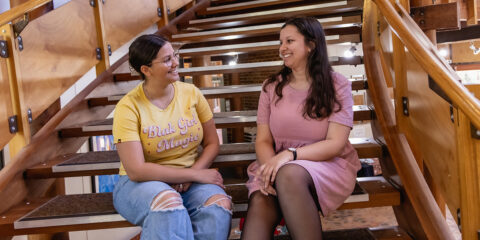 Two female indigenous students sitting on stairs and talking while smiling.