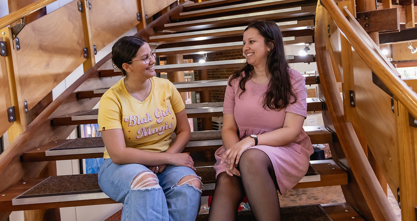 Two female indigenous students sitting on stairs and talking while smiling.