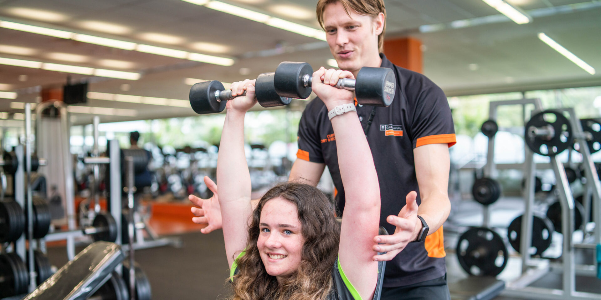 A female does a shoulder press with dumbbells, a Curtin Stadium staff member stands behind her.