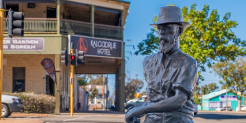 A bronze sculpture of a man wearing a hat sitting in front of the Kalgoorlie Hotel