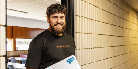 A student smiles at the camera holding a laptop.