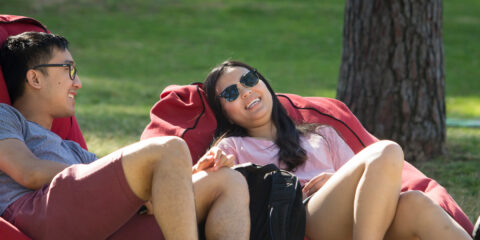 Two people relaxing on red bean bag chairs on a grassy campus lawn under a tree.